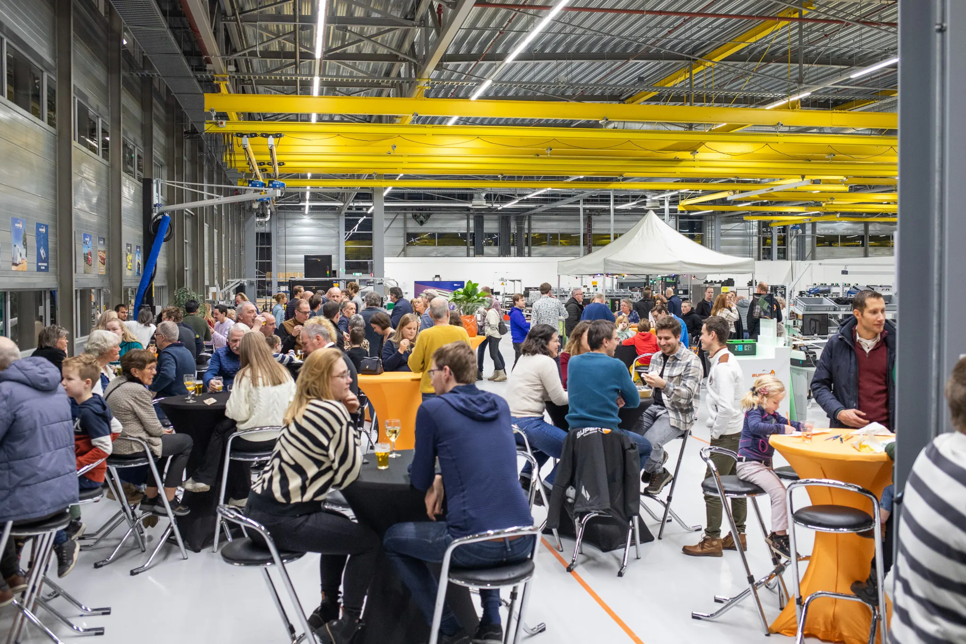 Mensen zitten aan tafels in een industriële Super B hal. Openingsfeest