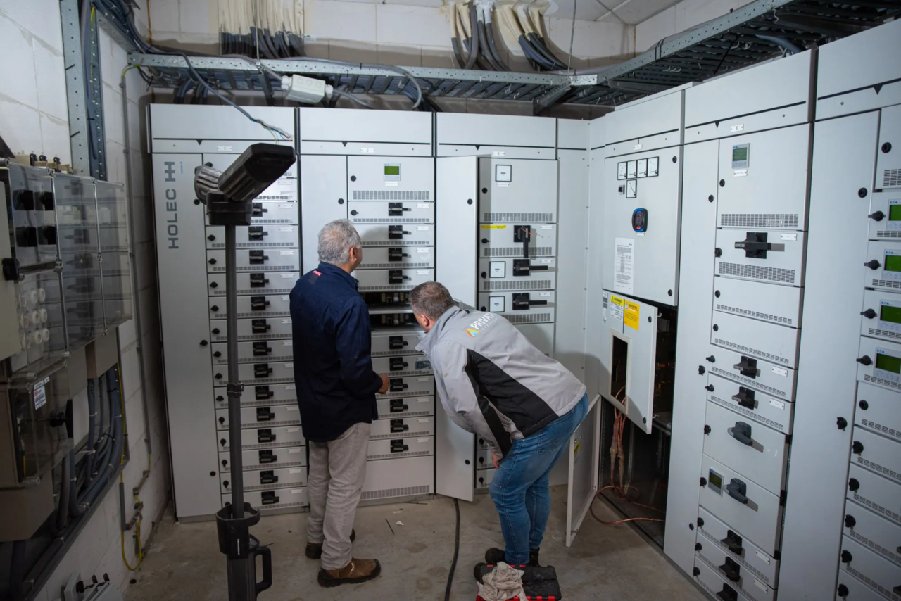Twee elektriciens werken aan een wit elektrisch schakelpaneel in een technische ruimte met kabels aan het plafond.