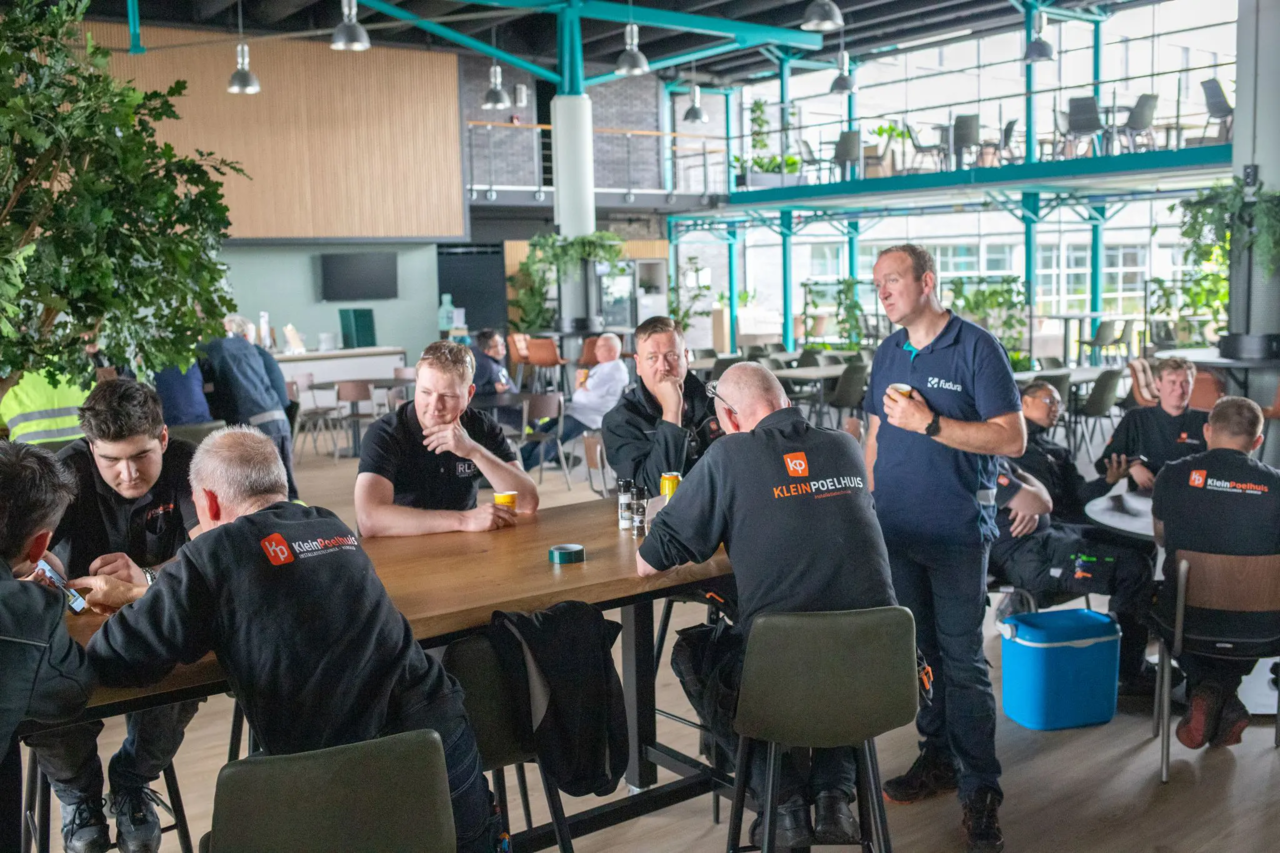 Werknemers van Klein Poelhuis in donkere werkkleding zitten samen aan tafels in de moderne bedrijfskantine tijdens pauze.