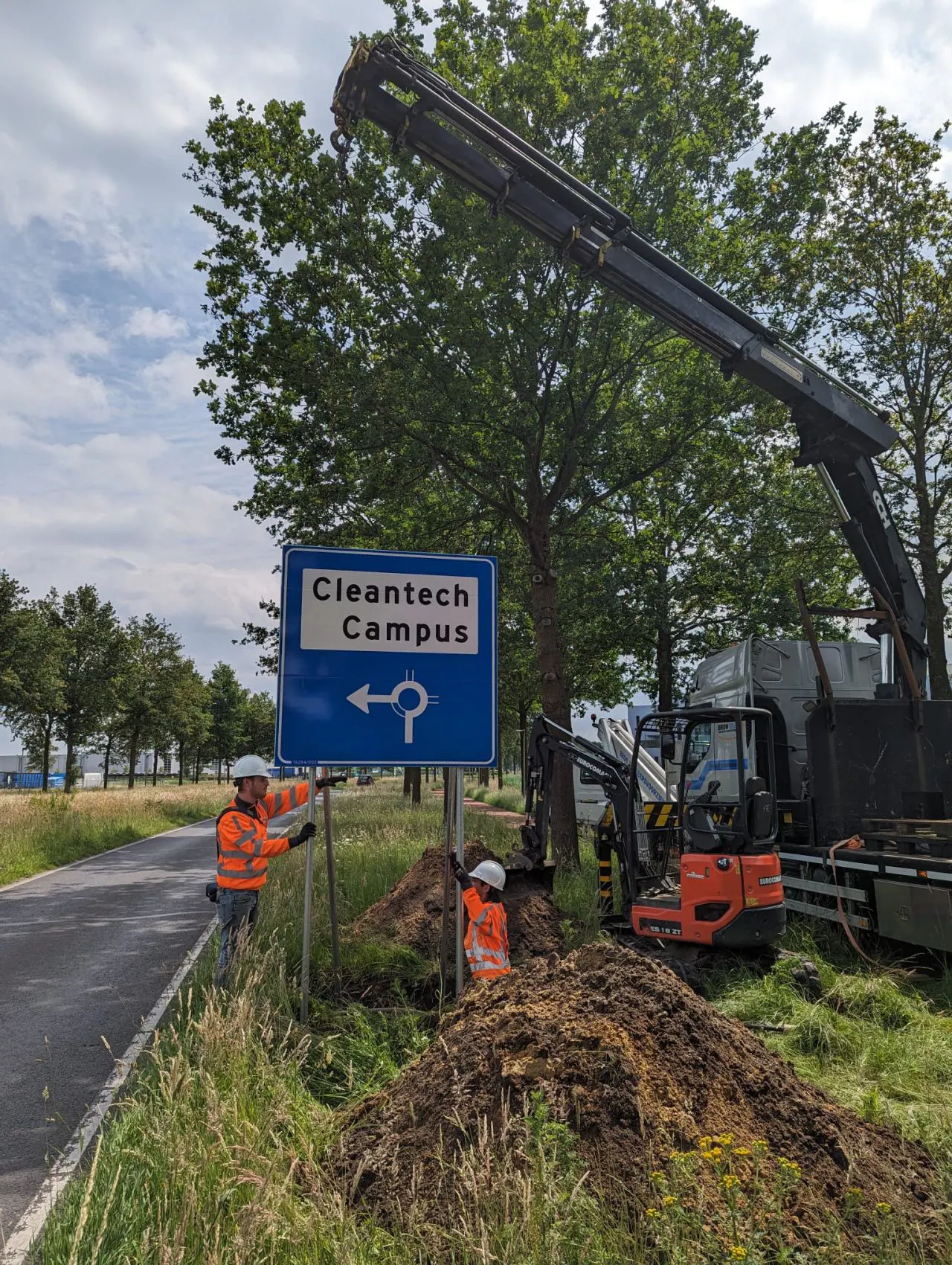 Werknemers installeren een blauw verkeersbord "Cleantech Campus" met graafmachine langs weg
