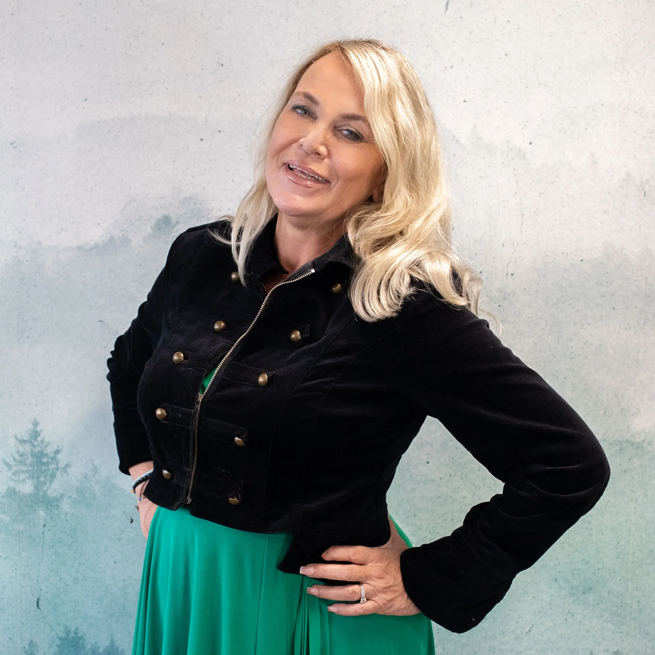 Blonde vrouw in zwarte militaire jas en groene rok poseert met hand op heup tegen witte muur