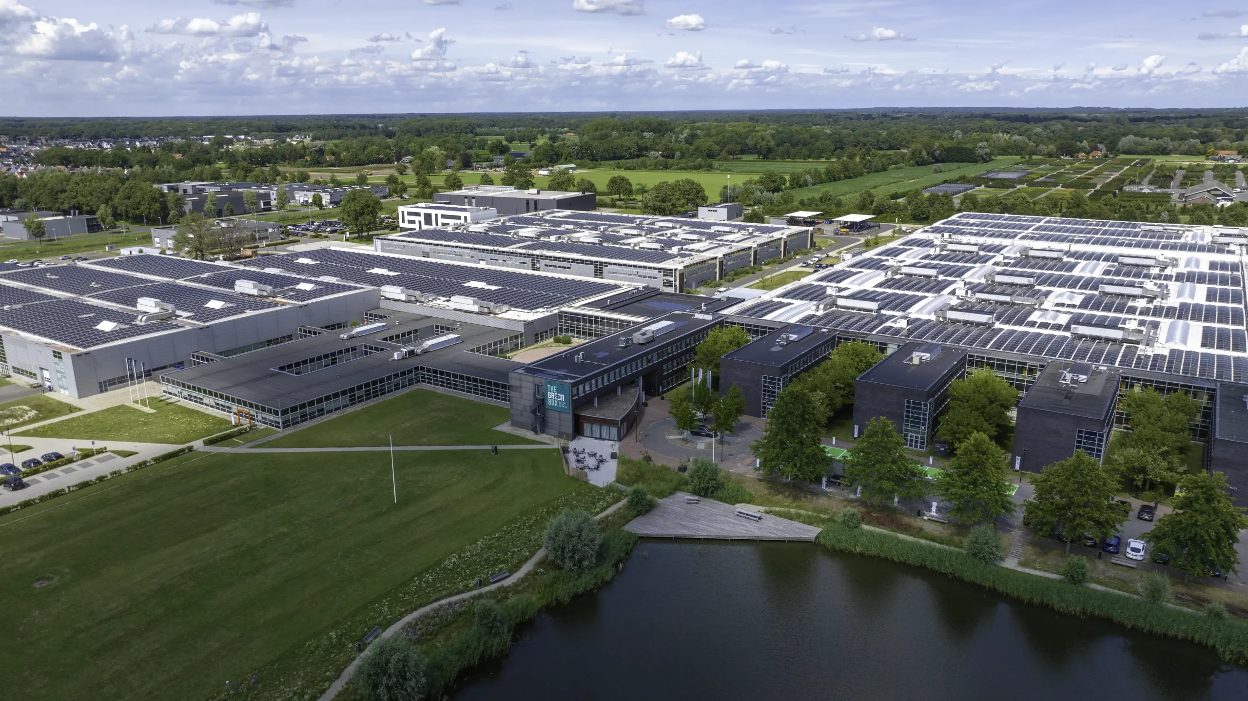 Luchtfoto van modern bedrijvencomplex met zonnepanelen op daken en water op de voorgrond, omgeven door groen landschap.