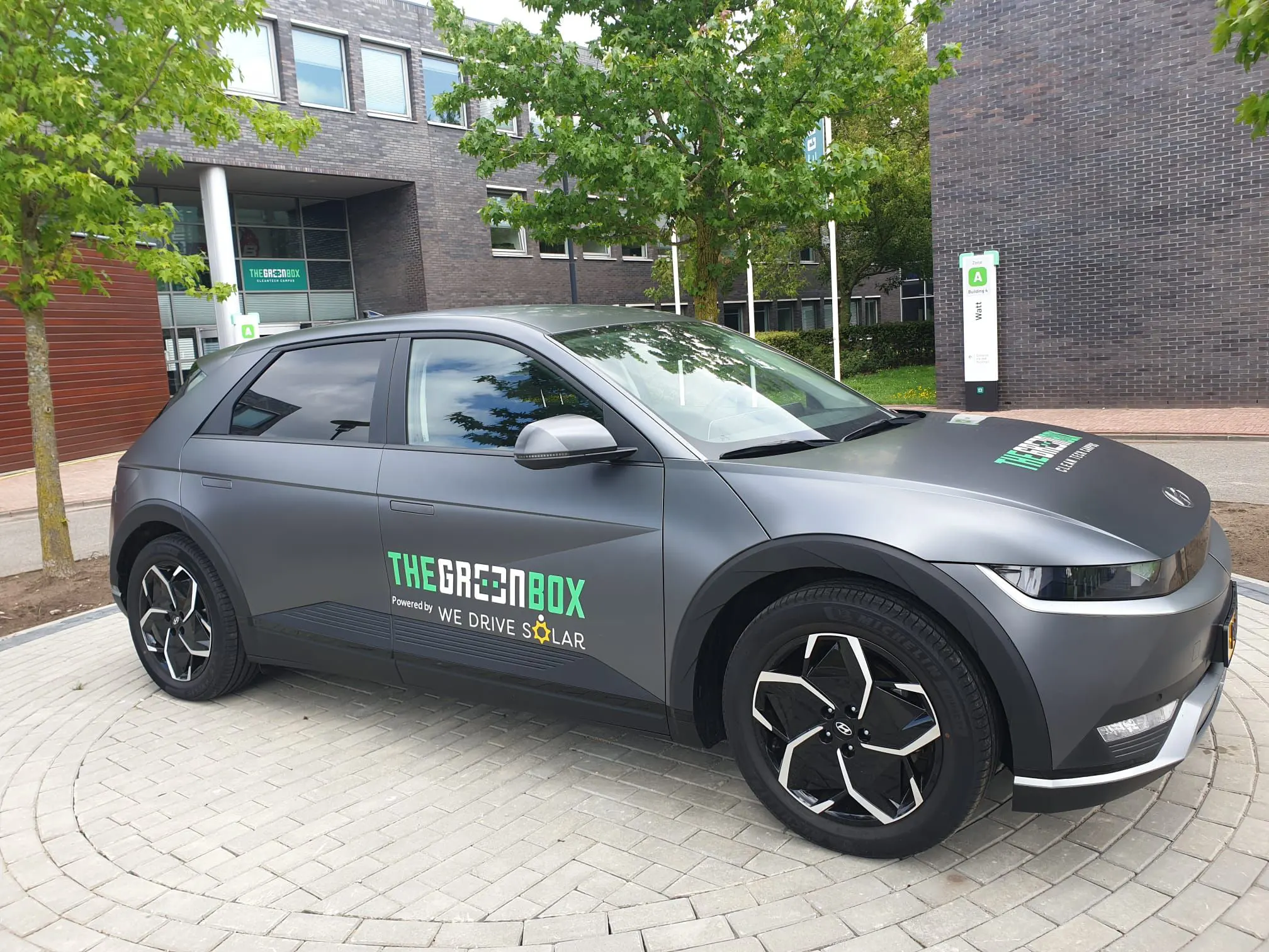 bidirectionele auto met "THEGREENBOX - WE DRIVE SOLAR" tekst geparkeerd bij laadstation