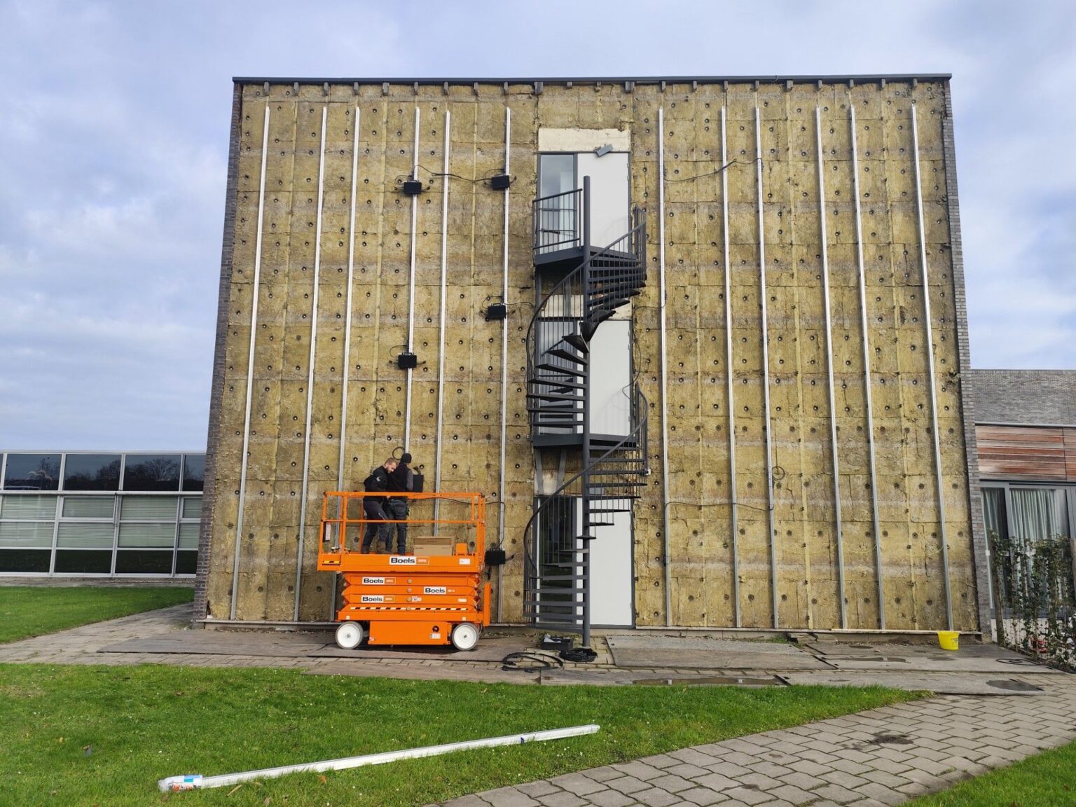 Arbeiders renoveren gevel van betonnen gebouw met oranje hoogwerker en externe wenteltrap
