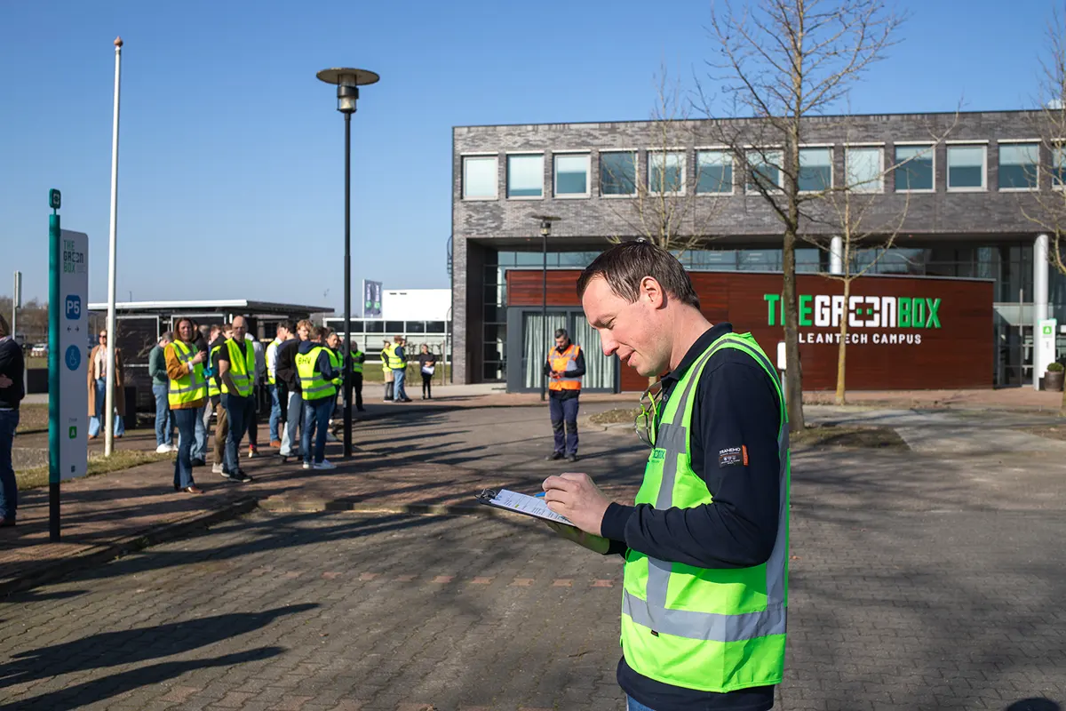 Man in veiligheidsvest met tablet op Greenbox Leantech Campus terwijl groep werknemers op achtergrond staat te wachten